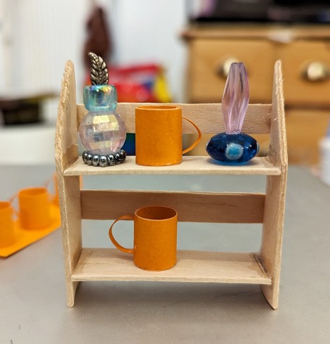 Miniature shelf from ice lolly sticks with 2 orange miniature cups out of paper and 2 flacons made out of big glass and plastic pearls and some metallic charms.